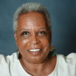 The image is a close-up portrait of Shakiri, a Black woman with short curly grey hair. She is smiling and has a warm, open expression. She’s wearing large earrings that are gold and jade green and an off-white tunic with buttons at her neckline. Photo by Nano Visser.