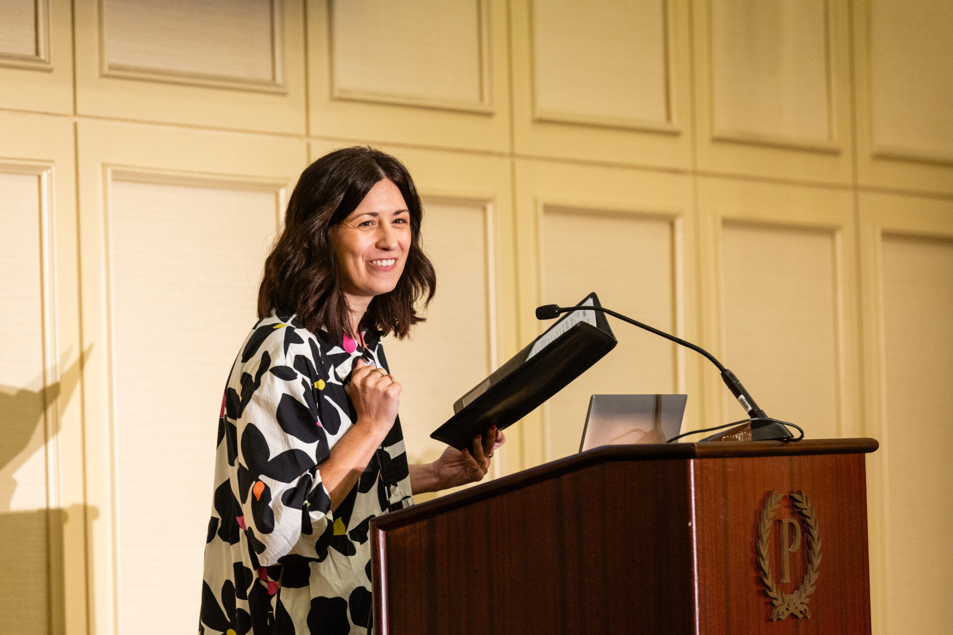 A woman with short dark hair smiling while speaking at a podium during a formal event, holding a folder and standing in front of a microphone. She wears a dress that's white with black flowers and pops of color.